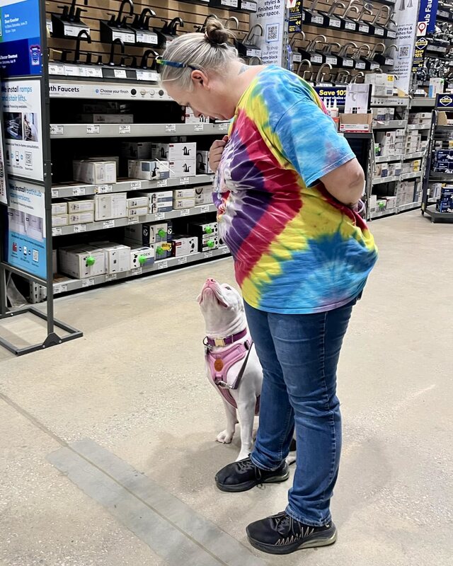 Wendy in a hardware store aisle smiling down at Penelope, a white pit bull in a pink harness, who sits and looks up at her