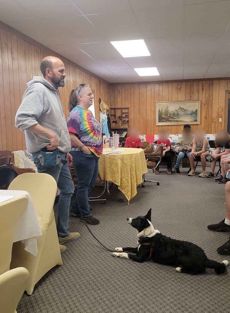 Will and Wendy standing at the front of a room talking to a homeschool group, with a black and white dog lying calmly at their feet